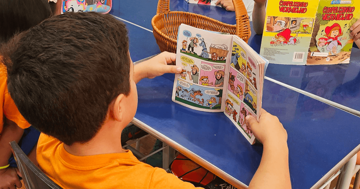 Criança lê uma história em quadrinhos sentada à mesa durante uma atividade escolar.